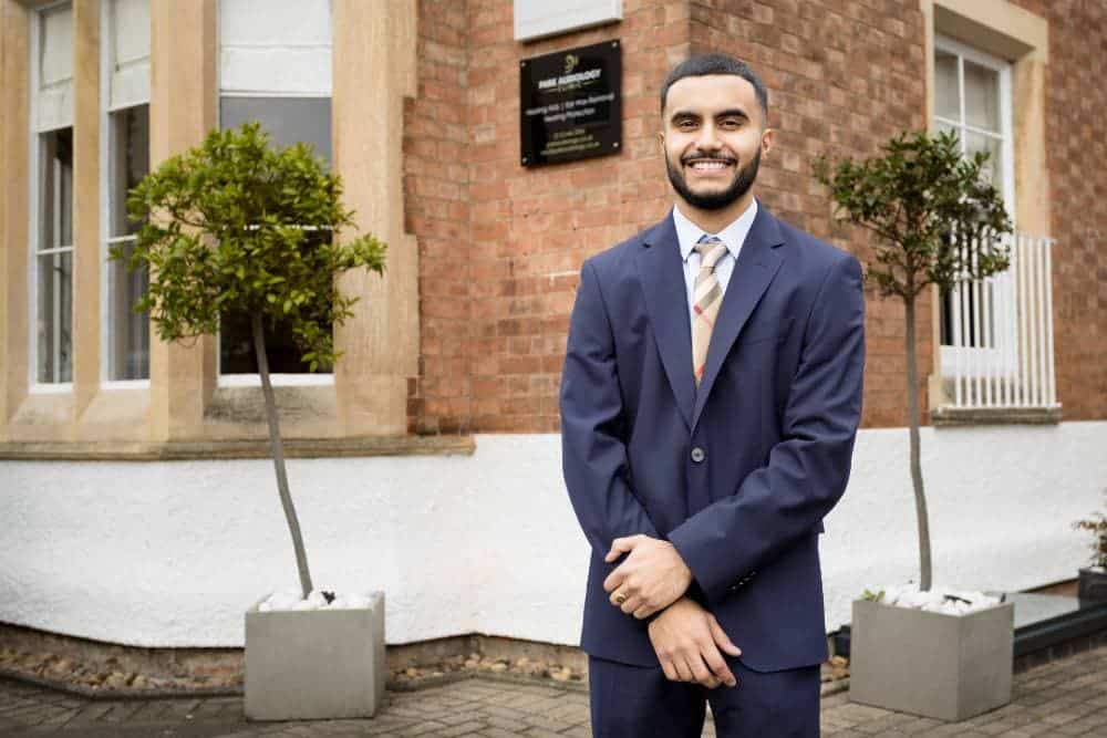 A man in a business suit smiling in front of a building with two potted plants.