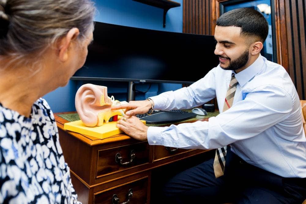 A male audiologist explaining a model of the human ear to an elderly female patient in a clinic.