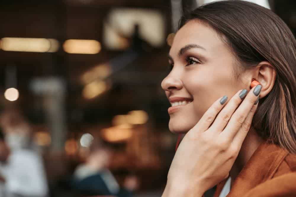 A woman smiling as she touches her wireless earpiece, viewed in profile inside a café.