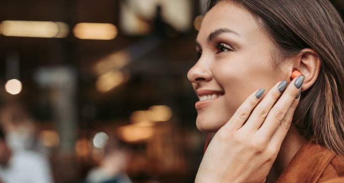 A side profile of a smiling woman with long hair, touching a wireless earbud while sitting in a café.